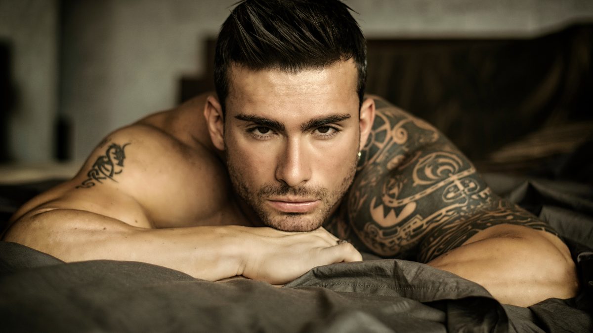 Shirtless male creator with a tattoo resting on a bed